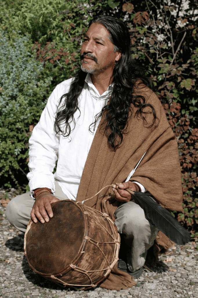 Man with long hair playing a traditional drum outdoors.
