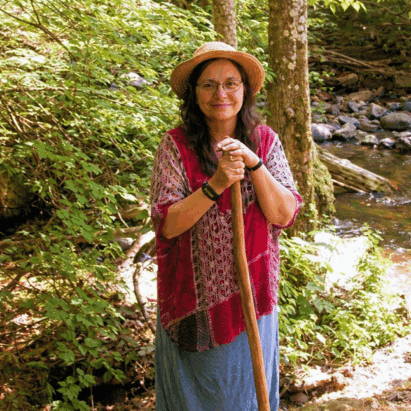 Woman in a hat and shawl standing with a walking stick in a forest.