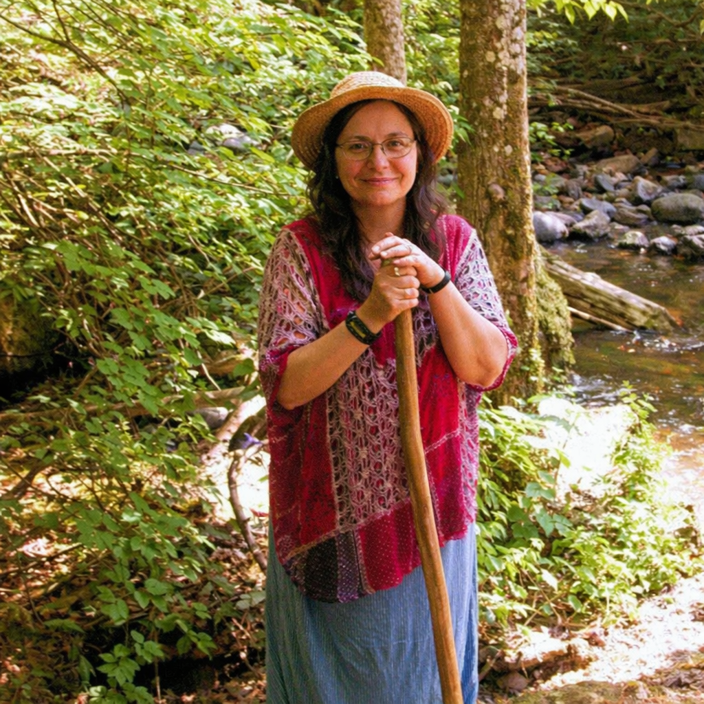 Woman in a hat and shawl standing with a walking stick in a forest.